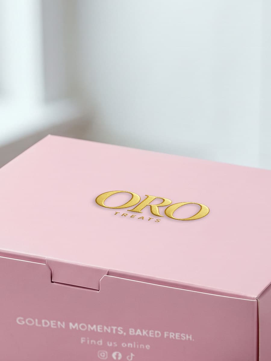A closed pink Oro Treats box, the gold wordmark on its lid and Golden moments, baked fresh printed on its front.