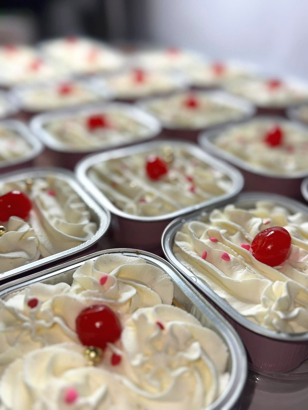 Finished Signature tres leches in their foil trays, piped with whipped cream and topped with a red cherry, pink sugar hearts and gold pearls.