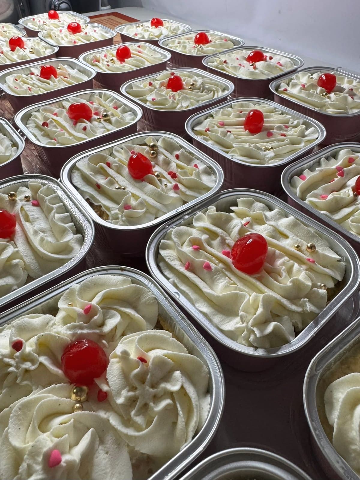 Rows of Signature tres leches in foil trays, each topped with whipped cream, a red cherry, pink sugar hearts and a gold pearl.
