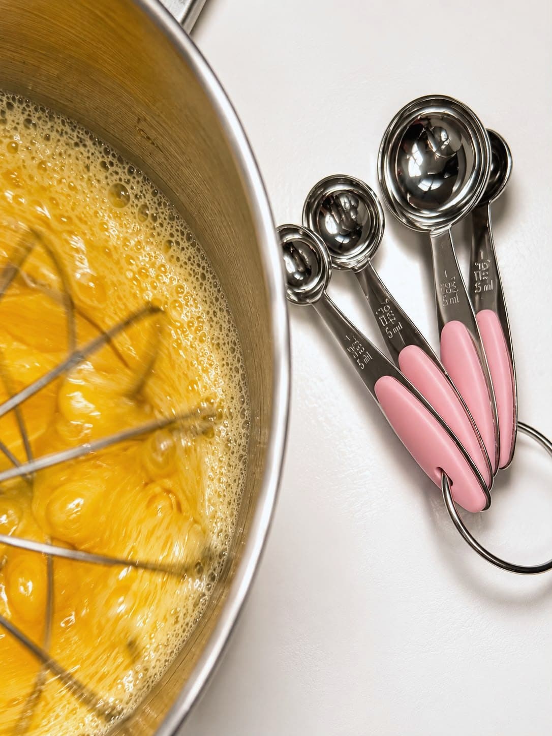 Eggs whisked in a steel mixer bowl, beside a set of pink-handled measuring spoons.